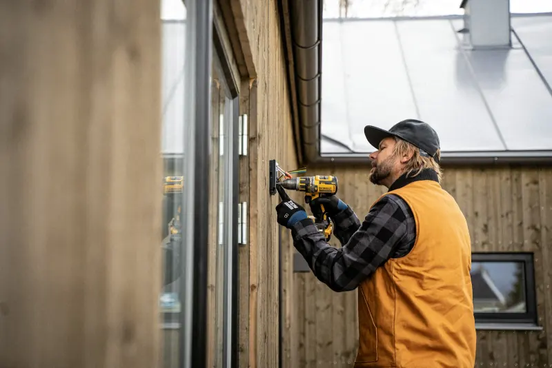 Man met een accuboormachine boort een gat in een houten gevel voor buitenverlichting
